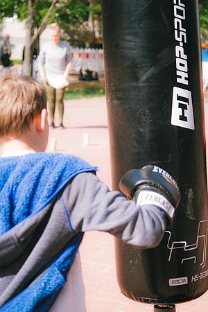 Ein Kind beim Boxtraining an einem schwarzen Boxsack im Freien. Sichtbar ist das Kind von hinten, es trägt Boxhandschuhe. Im Hintergrund eine unkenntliche Person und Bäume.
