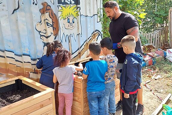 Kinder und ein Erwachsener befüllen hölzerne Hochbeete vor einer bemalten Container-Wand.