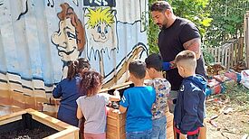 Kinder und ein Erwachsener befüllen hölzerne Hochbeete vor einer bemalten Container-Wand.