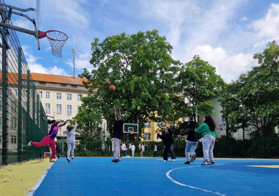 Jugendliche spielen auf einem eingezäunten blauen Sportplatz Basketball an zwei Körben . Im Hintergrund sind Wohngebäude und Bäume zu sehen.