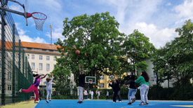 Jugendliche spielen auf einem eingezäunten blauen Sportplatz Basketball an zwei Körben . Im Hintergrund sind Wohngebäude und Bäume zu sehen.
