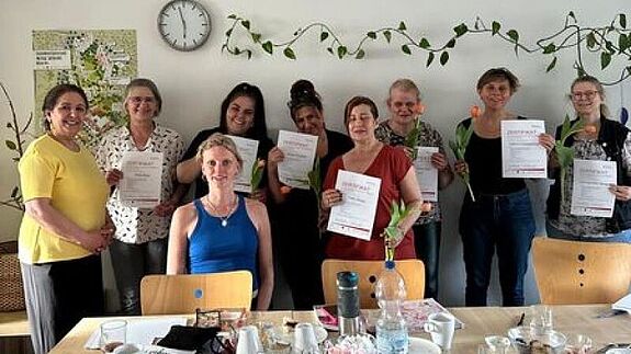 Neun Frauen stehen hinter einem gedeckten Tisch und halten Zertifikate in die Kamera.