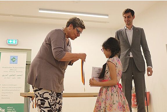Bildungsstadträtin Karin Korte übergibt eine Medaille an eine Schülerin. Im Hintergrund steht Bezirksbürgermeister Martin Hikel.. 