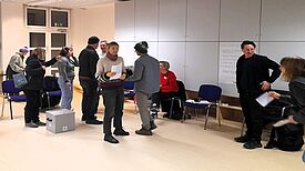Menschen stehen in einem Raum und unterhalten sich während der Quartiersratssitzung, einige halten Papiere in der Hand.
