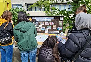 Eine Gruppe: Vier Erwachsene und ein Kind, von hinten sichtbar, stehen vor einem Tisch. Auf dem Tisch befindet sich eine Karte. Hinter dem Tisch seht eine Tafel mit Fotos und Text: „Mehr Freizeit- und Bewegungsorte für Erwachsene im Quartier“ und „Was bewegt dich?“. Im Hintergrund ist eine Hausfassade. 