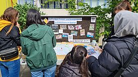 Eine Gruppe: Vier Erwachsene und ein Kind, von hinten sichtbar, stehen vor einem Tisch. Auf dem Tisch befindet sich eine Karte. Hinter dem Tisch seht eine Tafel mit Fotos und Text: „Mehr Freizeit- und Bewegungsorte für Erwachsene im Quartier“ und „Was bewegt dich?“. Im Hintergrund ist eine Hausfassade. 