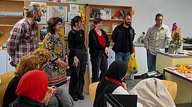 Mehrere Erwachsene und Jugendliche stehen in einem Raum der Boddinbox zusammen; einige sitzen, andere stehen, im Hintergrund sind Infoplakate, Regale mit Materialien und frisches Obst auf einem Tisch zu sehen.