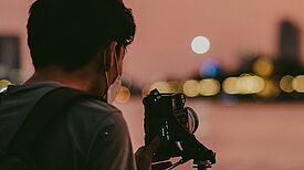 Eine Person fotografiert mit einer Kamera auf einem Stativ den Abendhimmel über einer unscharfen Stadtansicht.