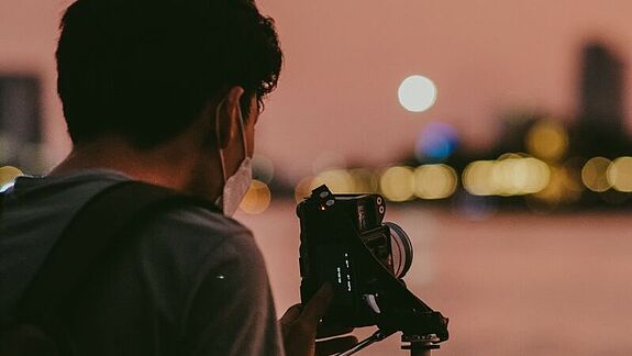 Eine Person fotografiert mit einer Kamera auf einem Stativ den Abendhimmel über einer unscharfen Stadtansicht.
