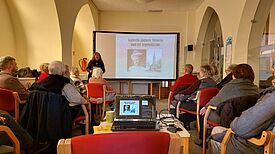 Eine Gruppe Menschen sitzt vor einer Leinwand, auf die der Lichtbildvortrag zum Leben der Kaiserin Auguste Viktoria projiziert wird.