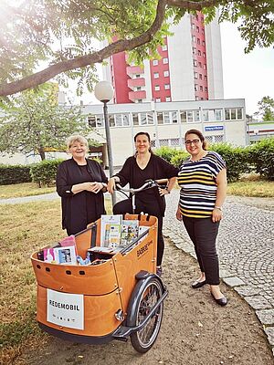 Drei Frauen stehen lächelnd hinter einem Lastenrad unter einem Baum. Auf dem Lastenrad steht „Redemobil“, es ist mit farbigen Broschüren gefüllt. Im Hintergrund ein einstöckiger Funktionsbau und ein Wohnhochhaus 
