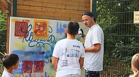 Ein Erwachsener zeigt drei jungen Teilnehmenden an einer mit Schablonen und Sprühdosen gestalteten im Freien Wand Graffiti-Techniken.