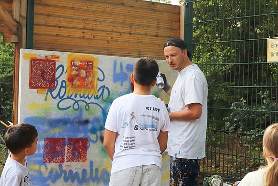 Ein Erwachsener zeigt drei jungen Teilnehmenden an einer mit Schablonen und Sprühdosen gestalteten im Freien Wand Graffiti-Techniken.