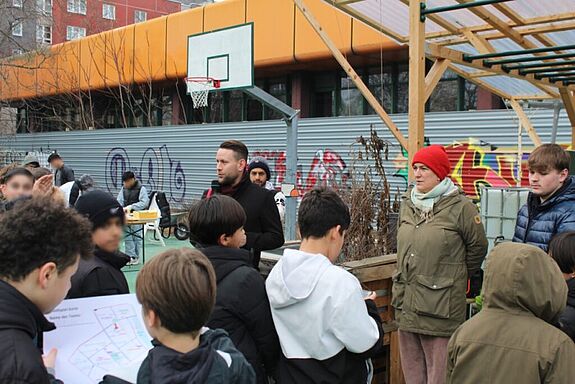 Jugendliche und Anwohnende stehen während des Kiezfests auf einem ehemaligen Sportplatz, umgeben von Hochbeeten, Pavillons und einem Basketballkorb.