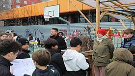 Jugendliche und Anwohnende stehen während des Kiezfests auf einem ehemaligen Sportplatz, umgeben von Hochbeeten, Pavillons und einem Basketballkorb.