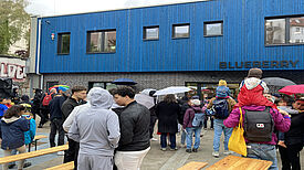 Vor einem blauen Gebäude stehen Erwachsene, Jugendliche und Kinder, die sich locker unterhalten. Vor dem Gebäude sind Tische und Bänke aufgebaut.  Auf dem Gebäude steht „BLUEBERRY“.