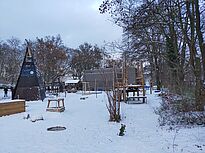 Die verschneite Außenanlage zeigt im Vordergrund Spielgeräte und Kletterelemente zwischen kahlen Bäumen. Im Hintergrund steht der Neubau des Moabiter Kinderhofs, der von der winterlichen Parklandschaft umgeben ist.