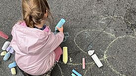 Ein Kind malt mit bunter Straßenkreide auf den Asphalt einer gesperrten Straße, umgeben von großen Kreidestücken in verschiedenen Farben.