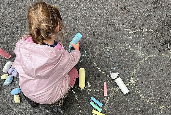 Ein Kind malt mit bunter Straßenkreide auf den Asphalt einer gesperrten Straße, umgeben von großen Kreidestücken in verschiedenen Farben.