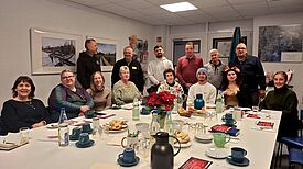 Menschen sitzen und stehen um einen mit Blumen dekorierten, gedeckten Tisch.