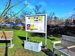 Zwei Sitzbänke aus Holz und Metall auf einem Rasenstück, zwischen ihnen eine Infotafel und ein bepflanzter Kübel. Im Hintergrund Sträucher, ein Parkplatz und Wohnhochhäuser.