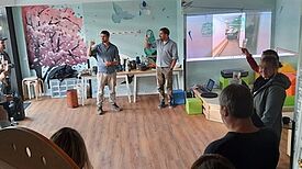 Menschen stehen in einem freundlich gestrichenen Raum mit Wandillustrationen und erheben ihre Gläser.