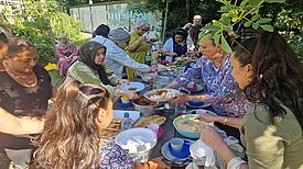Menschen aus der Nachbarschaft treffen sich an einer langen Tafel im Gemeinschaftsgarten Prinzessinengärten und essen gemeinsam die gekochten Speisen.
