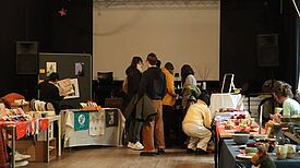 Mehrere Menschen stehen in einem Raum, in dem es verschiedene Kunstmärkte zu sehen gibt. 