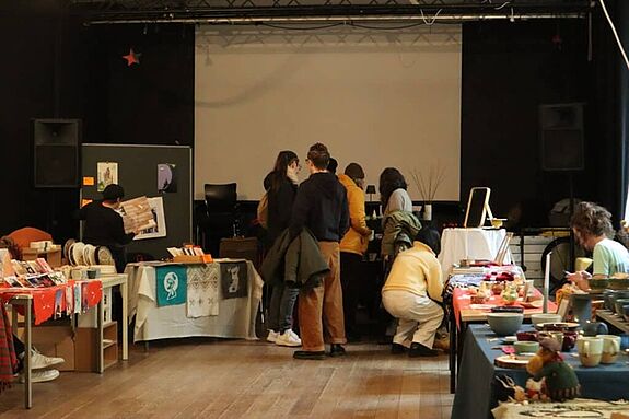 Mehrere Menschen stehen in einem Raum, in dem es verschiedene Kunstmärkte zu sehen gibt. 