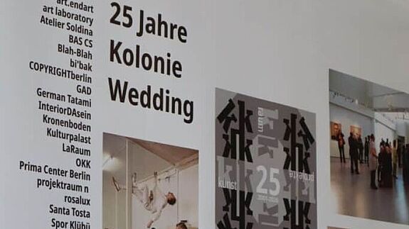 Ausstellungstafel zu 25 Jahren Kolonie Wedding mit Fotos, Plakaten und Texten in einem Projektraum.