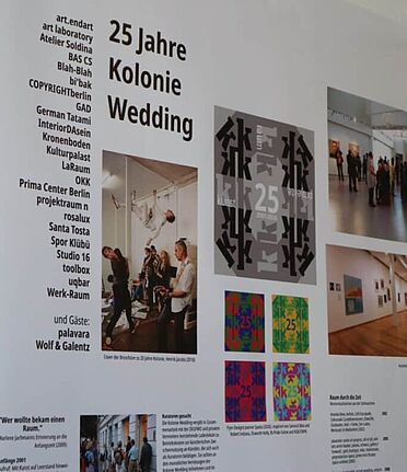 Ausstellungstafel zu 25 Jahren Kolonie Wedding mit Fotos, Plakaten und Texten in einem Projektraum.