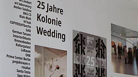 Ausstellungstafel zu 25 Jahren Kolonie Wedding mit Fotos, Plakaten und Texten in einem Projektraum.