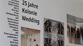 Ausstellungstafel zu 25 Jahren Kolonie Wedding mit Fotos, Plakaten und Texten in einem Projektraum.