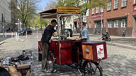 Ein umgebautes Lastenrad steht auf einer gepflasterten Straße. Zwei Männer stehen an dem Rad und unterhalten sich miteinander. 