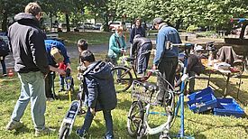 Erwachsene und Kinder reparieren gemeinsam Fahrräder und Roller im Freien bei einer Mitmachaktion auf einer Wiese.