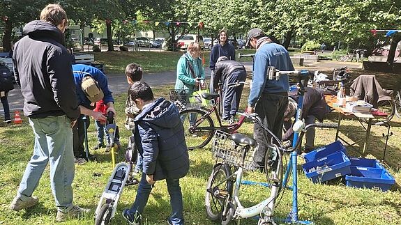Erwachsene und Kinder reparieren gemeinsam Fahrräder und Roller im Freien bei einer Mitmachaktion auf einer Wiese.