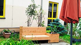 Ein Hochbeet aus hellem Holz mit integrierter Sitzbank steht im begrünten Innenhof vor einem Gebäude mit gelben Fensterrahmen.