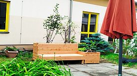 Ein Hochbeet aus hellem Holz mit integrierter Sitzbank steht im begrünten Innenhof vor einem Gebäude mit gelben Fensterrahmen.