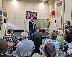 Der Staatssekretär für Wohnen und Mieterschutz, Stephan Machulik, eröffnet die Veranstaltung. Er steht im Innenraum des Neubaus. Vor ihm sitzen Gäste und hören seinem Grußwort zu. Im Zentrum des Vortrags steht der Wert des Moabiter Kinderhofes für den Kiez sowie der Dank an die vielen Mitwirkenden des Förderprojektes.