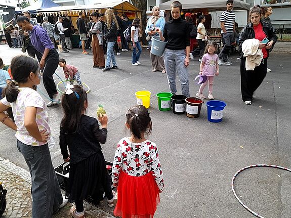 Auf einem Straßenfest werfen Kinder Dinge in fünf verschiedenfarbige, beschriftete Eimer. 