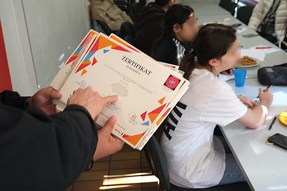 Eine Person hält mehrere Zertifikate für die „Kiezheld*innen“-Ausbildung, während Jugendliche am Tisch sitzen und aufmerksam am Workshop teilnehmen.