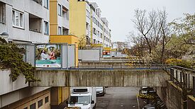 Wohnungsneubauten mit weiß und gelb gemalter Fassade. Eine Betonüberführung führt von bepflanzten Rabatten zum Eingang. An einer Außenwand hängt ein buntes Plakat auf dem Menschen abgebildet sind.  