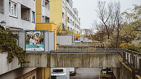 Wohnungsneubauten mit weiß und gelb gemalter Fassade. Eine Betonüberführung führt von bepflanzten Rabatten zum Eingang. An einer Außenwand hängt ein buntes Plakat auf dem Menschen abgebildet sind.  