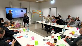 Die Mitglieder des Quartiersrats sitzen an Tischen während zwei Personen etwas präsentieren. 