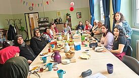Frauen sitzen gemeinsam an einer langen gedeckten Tafel in einem hellen Raum.