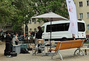 Erwachsene und Kinder bauen draußen auf einem Platz technische Geräte auf. Bei ihnen steht eine Bank, mehrere Kisten und Geräte, ein Sonnenschirm und eine Werbeflagge. Hinter ihnen ein weißer Kleinbus mit der Aufschrift „Kiezsportbus. Dein Kiez in Bewegung“. Im Hintergrund Bäume und Wohnhäuser.