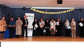 Mehrere Menschen stehen nebeneinander auf einer Bühne und halten ein Zertifikat in den Händen. 