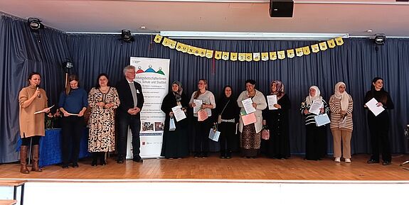 Mehrere Menschen stehen nebeneinander auf einer Bühne und halten ein Zertifikat in den Händen. 