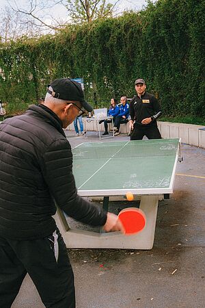 Tischtennisspiel im Freien, bei dem zwei Männer mit roten Schlägern und orangefarbenem Ball gegeneinander antreten. Im Hintergrund sitzen Zuschauer in Trainingsanzügen an einem Tisch, umgeben von Hecken und Bäumen.