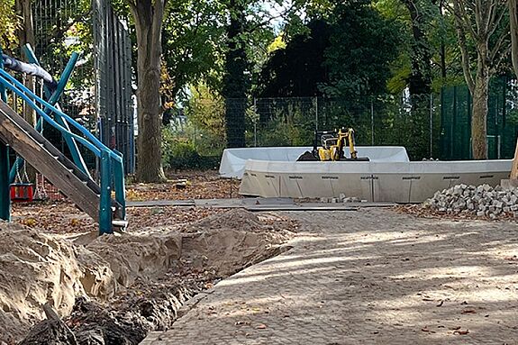 Ein kleiner Bagger arbeitet an einem Graben auf dem teilweise gepflasterten Spielplatz Silbersteinstraße.